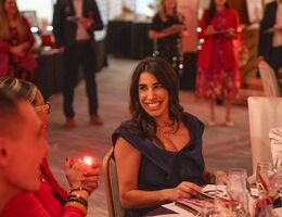 Guests sit at an elegantly set table during a gala event, surrounded by warm lighting, glassware, and other attendees in the background.