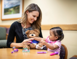 female child life specialists sits with patient who is playing doctor on a teddy-bear