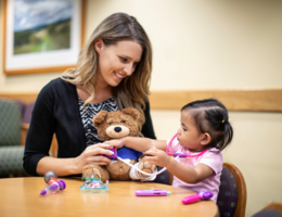 female child life specialists sits with patient who is playing doctor on a teddy-bear