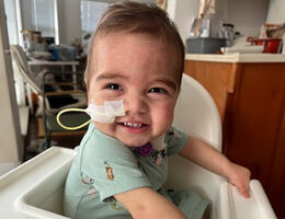 one year old boy sitting in high chair, smiling at camera, medical tube in nose