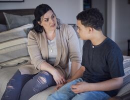 A Caucasian mother talking with mixed-race son while both sit on a bed
