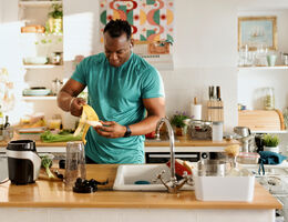 An African-American male in a bright kitchen prepares a banana at a wooden countertop surrounded by cooking tools, fresh produce, and a blender, with colorful artwork and neatly arranged shelves in the background.