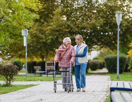Senior woman and her home caregiver spending a chilly, windy day outdoors in city park. Autumn walk for elderly patient with walker