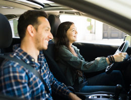 smiling father in passenger seat while teenage daughter sits in drivers seat with two hands on wheel, smiling