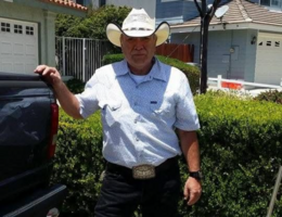A person wearing a white cowboy hat stands beside a pickup truck on a sunny residential street.