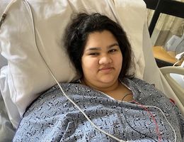 Teenge girl in hospital gown and hooked up to wires lies in bed and smiles at the camera