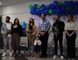 Six people stand shoulder to shoulder in front of blue and green balloon backdrop.