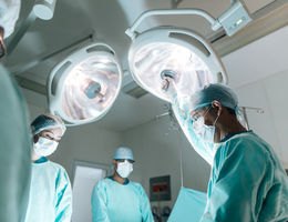 Four health workers wearing surgery clothes and masks, standing face to face in operation room with lamps on top.