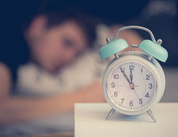 Alarm clock on nightstand in foreground, person sleeping in the background