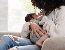 Sitting on a chair in her living room, the unrecognizable mom holds her baby snugly while breastfeeding him