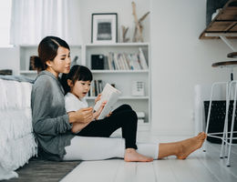 Young Asian mother sitting on the floor in the bedroom reading book to little daughter, enjoying family bonding time together at home