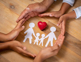 Close up hands of father, mother and daughters protecting family paper cutout with red heart. Hands of mixed race family with paper cutting and red heart shaped symbol stone on wooden background.