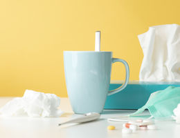 Tissue box, tissues, thermometer, protective face mask, capsules and a hot drink on white table in front of yellow wall.