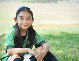 elementary age Hispanic female soccer player sits on the ground after her game. She is smiling big and holding a soccer ball. She has long brown hair and brown eyes. The playing field and soccer goal are in the background.