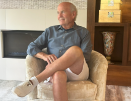 White man in a blue button up shirt and white shorts sits in a yellow chair in front of a fireplace