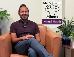 A man in a purple button up shirt sits in a cushioned orange chair in a therapist office. There are plants on side tables
