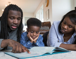 A preschool-age African American boy squeezes between his parents for reading time. They are sitting and sprawled out in the living room. He is resting his chin in both hands.