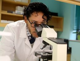 A doctor in a white lab coat and black hair looks through a microscope