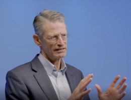 a man in front of a blue background is talking with his hands up 