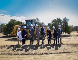 eight hospital leadership representatives dig dirt during the groundbreaking ceremony in front of a dump truck with trees in the background.