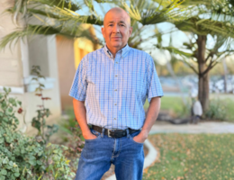Man in blue button up and jeans stands in front of his house with his hands in his pocket