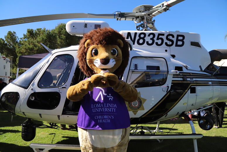 Loma Linda University Children's Hospital mascot Luke the Lion stands in front of helicopter and poses with heart hands