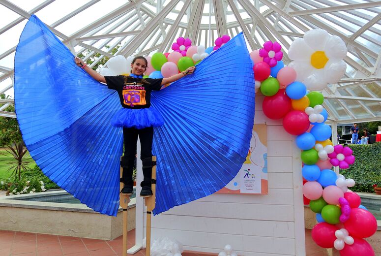 A performer stands elevated while holding large blue fabric wings in front of a colorful balloon display.