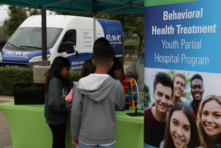 Children visit a behavioral health information booth under a canopy tent.