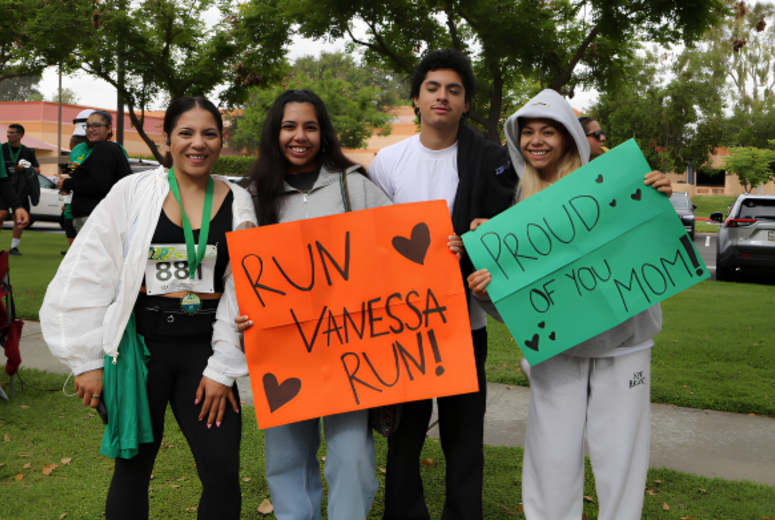 Participants hold supportive signs reading “Run Vanessa Run!” and “Proud of You Mom!” at a 5K event.