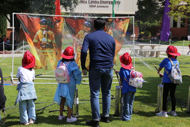 Children wearing red firefighter helmets spray water hoses at a target during a hands‑on fire safety activity.