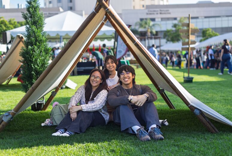 three students sitting on the lawn under a teepee style tent on the campus lawn