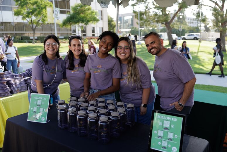 five students in matching purple t-shirts posing behind a table, smiling at the camera at welcome back bash