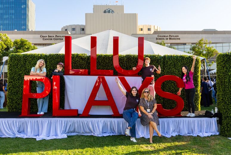 big red block letters that say "LLU PLAYS: on a stage with a smiling group of people posed around it