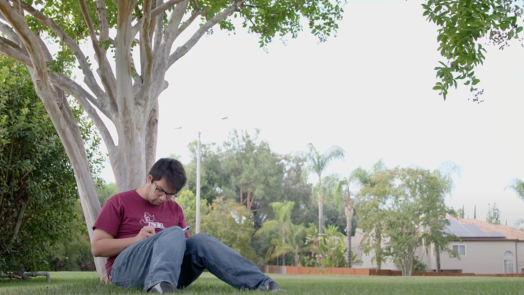 Tyler Rojanaroj sits at the base of a tree writing music.