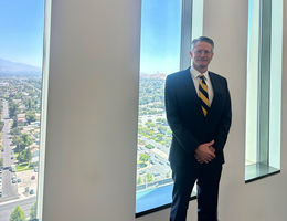 Caucasian man standing in a blue suit with blue and yellow striped tie in front of a window smiling with hands folded in front of him.