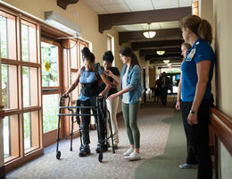 Black woman uses an assisted walking device while receiving physical therapy with the help of two other women in a hallway next to a window.