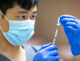 Asian male in blue scrubs, gloves, and surgical mask fills vaccine syringe.