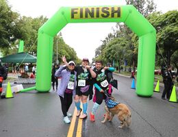 Three female participants and a dog stand in front of the green finish line holding their race medals