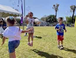 Three children in tee-shirts and shorts play outside in the grass blowing bubbles and smiling 