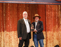 tall Caucasian man with white hair in a suit accepting award on stage from another Caucasian man in a suit and cowboy hat.