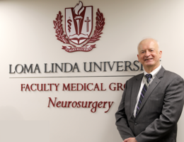 Man stands in front of a white wall with a sign saying Loma Linda University Faculty Medical Group Neurosurgery