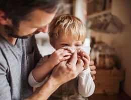 A father on paternity leave looking after small son indoors, blowing his nose