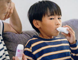 Boy using inhaler while sitting on gray couch, mom next to him supervising