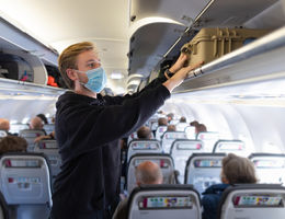A young backpacker storing his luggage away while on a plane, budget travelling. He is wearing a protective face mask to reduce the spread of COVID-19.