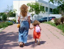 Woman with curly hair holds hands with a child with curly hair and a backpack as they walk toward a school
