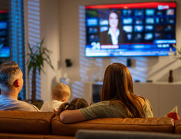 A woman, man, and baby sit on a brown couch and watch the news