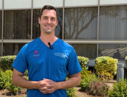 Doctor in blue scrubs stands outside a building surrounded by green bushes