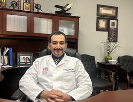 Man sits at desk smiling in white doctors coat.