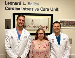 Colleen Barber reunites with the structural interventional cardiologists who recently performed a procedure to close a hole in her heart, Dr. Jason Hoff (left) and Dr. Amr Mohsen (right), framed by a background of photographs of Dr. Leonard Bailey who transplanted Barber's heart when she was a baby.
