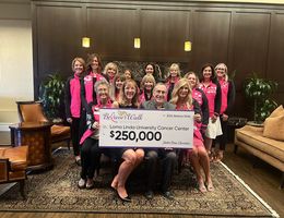 Group of women in pink jackets surrounding woman and man sitting holding oversized check reading a $250,000 donation to the Believe Walk